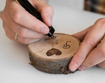 A close up image of a pair of hands burning a heart and other shapes into a wood coin using a wood burning tool
