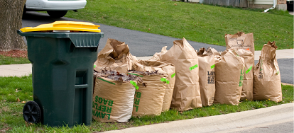 A row of stickered lawn bags next to a wheeled garbage tote